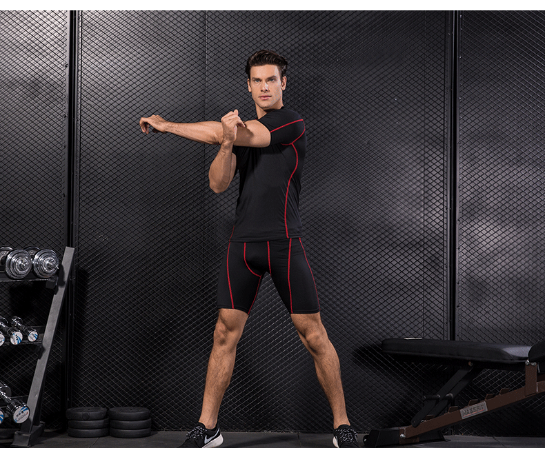 Man wearing black compression gym T-shirt and shorts with red accents performing physical exercises in a fitness studio.