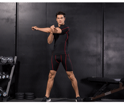 Man wearing black compression gym T-shirt and shorts with red accents performing physical exercises in a fitness studio.