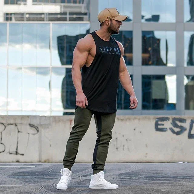 Muscular man wearing a black tank top with text, green pants, and white sneakers, standing in an urban setting.