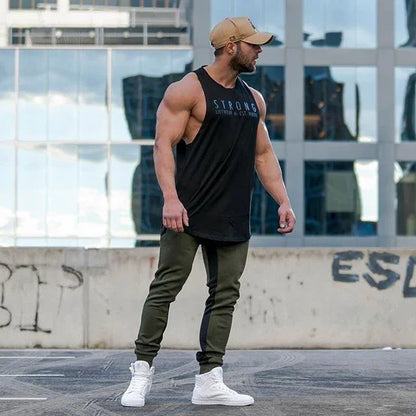 Muscular man wearing a black tank top with text, green pants, and white sneakers, standing in an urban setting.