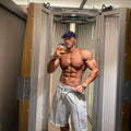 Muscular man taking a mirror selfie (hair removal chamber) wearing gray shorts and a cap.