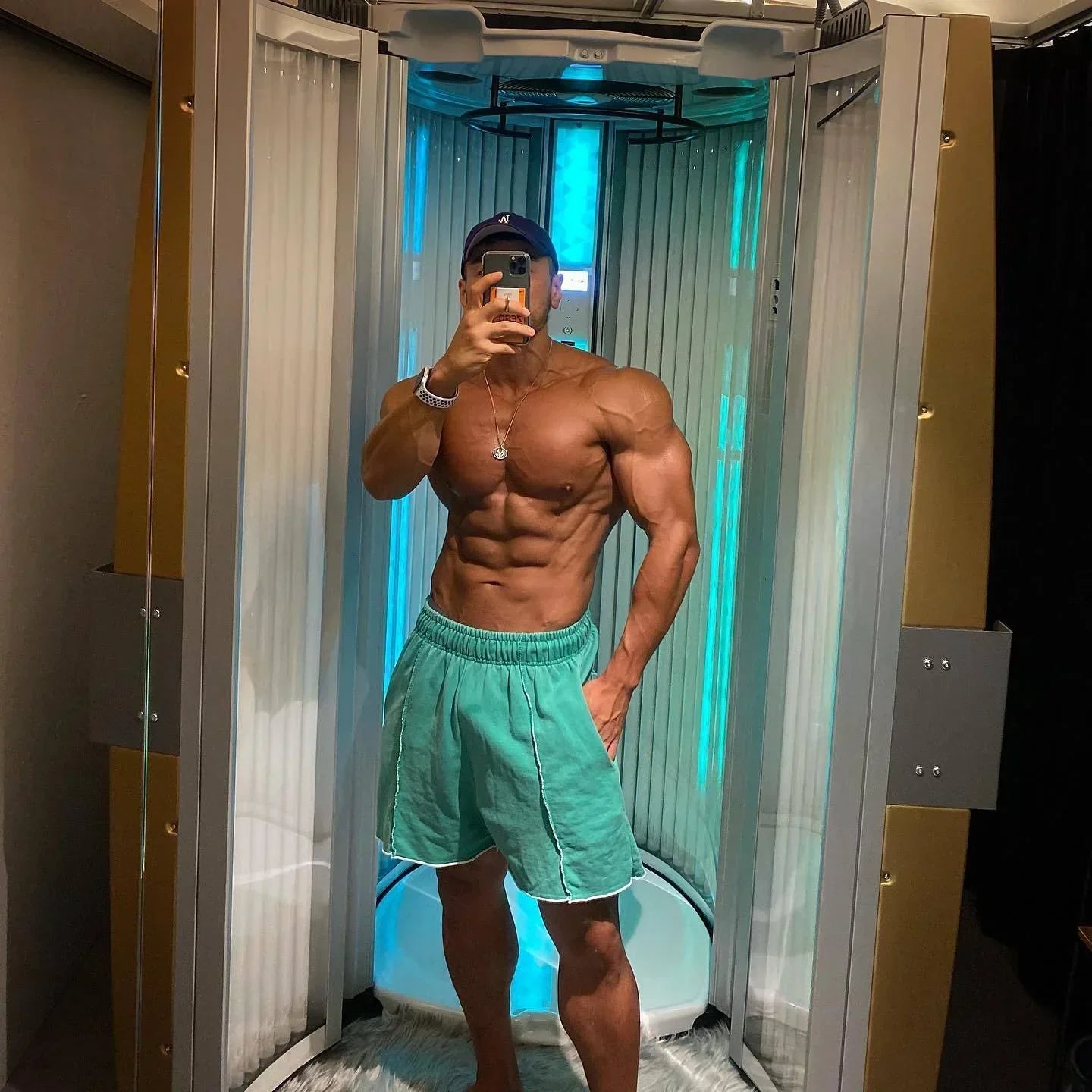 Man taking a mirror selfie in a tanning booth wearing teal shorts.