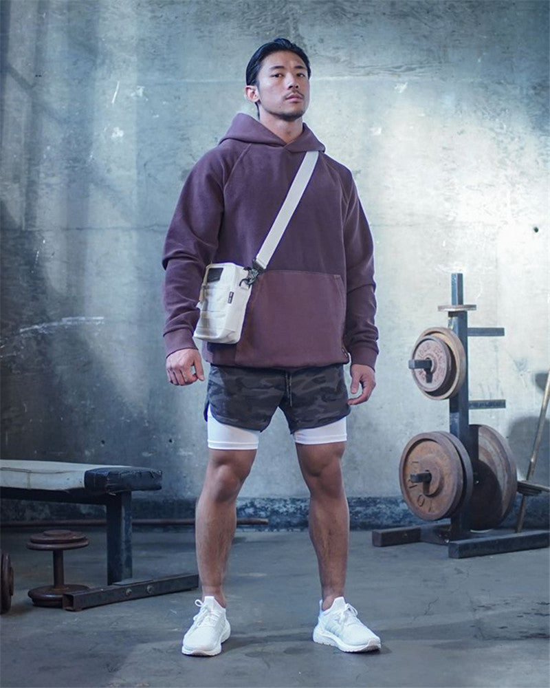 Man in a gym wearing a hoodie and shorts with a crossbody bag.