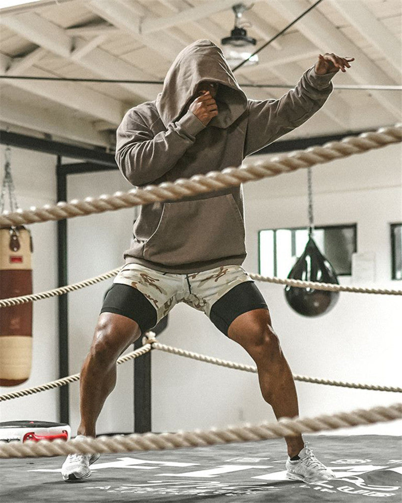 Person in a gray hoodie and shorts standing in a boxing ring with ropes.