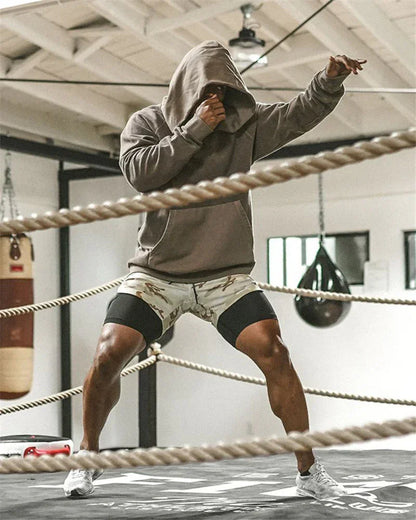 Person in a gray hoodie and shorts standing in a boxing ring with ropes.