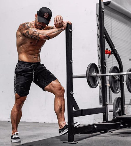 Muscular man in black shorts and cap exercising with weights in a gym.