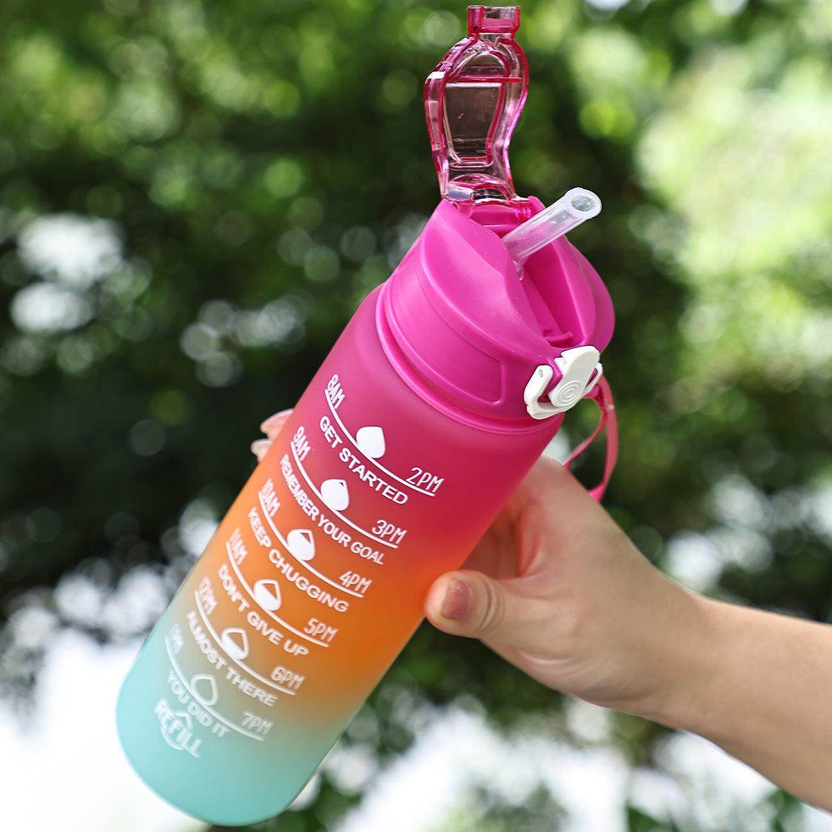 Gradient pink to blue water bottle with a straw and clip, held in a hand with a blurred green background.