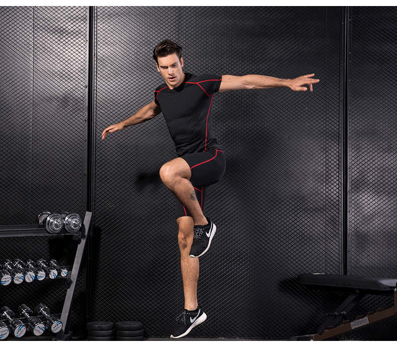 Man wearing black compression gym T-shirt and shorts with red accents standing in one leg in a fitness studio.