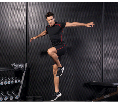 Man wearing black compression gym T-shirt and shorts with red accents standing in one leg in a fitness studio.