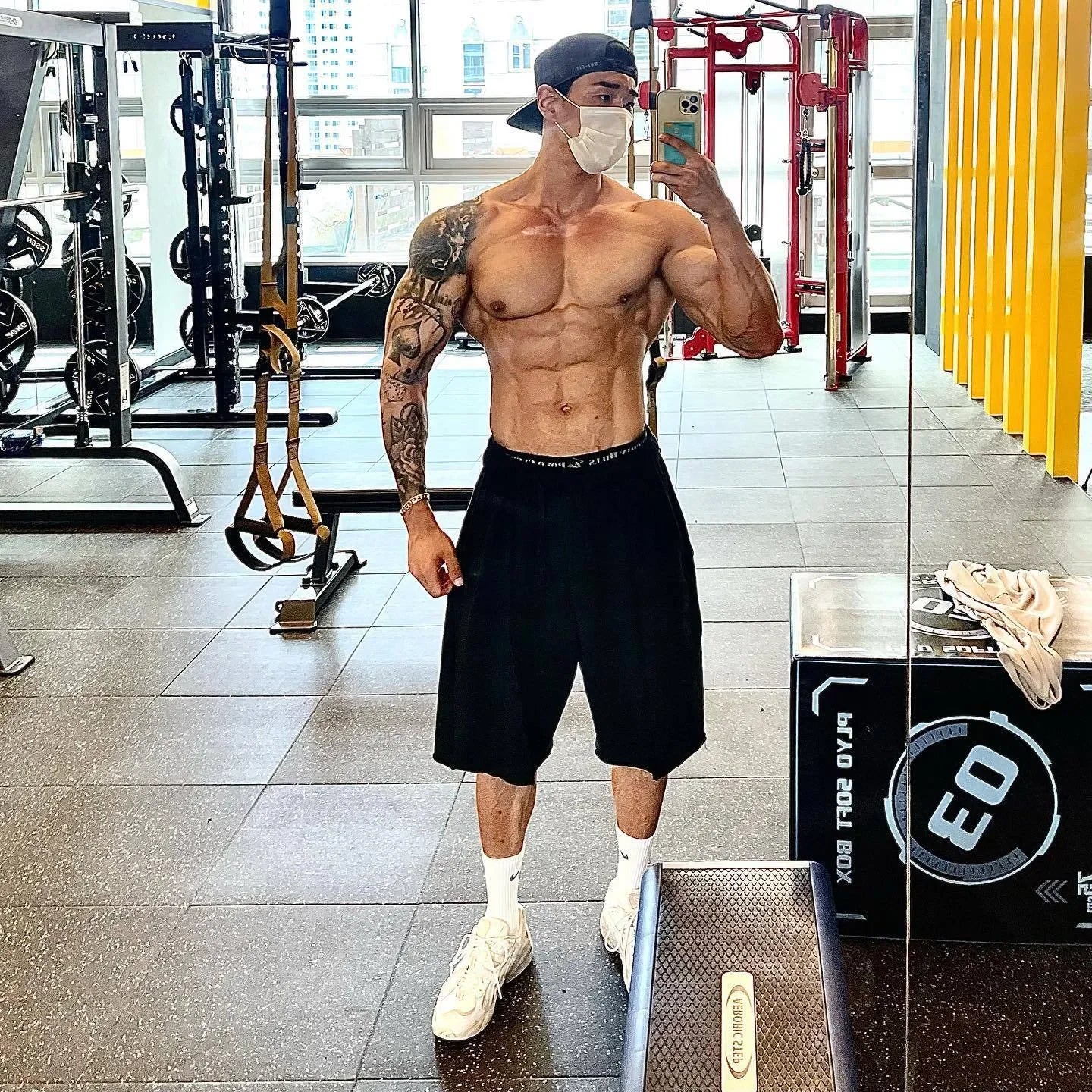 Muscular man in a gym wearing a face mask, black shorts, and white sneakers.