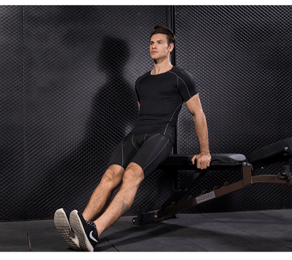 Man wearing black compression gym T-shirt and shorts with grey accents performing tricep dips on a bench in a fitness studio.